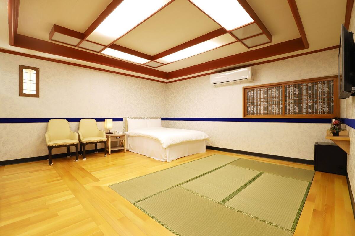 A hotel room with a bed, two chairs, a small table, a TV, and an air conditioning unit.