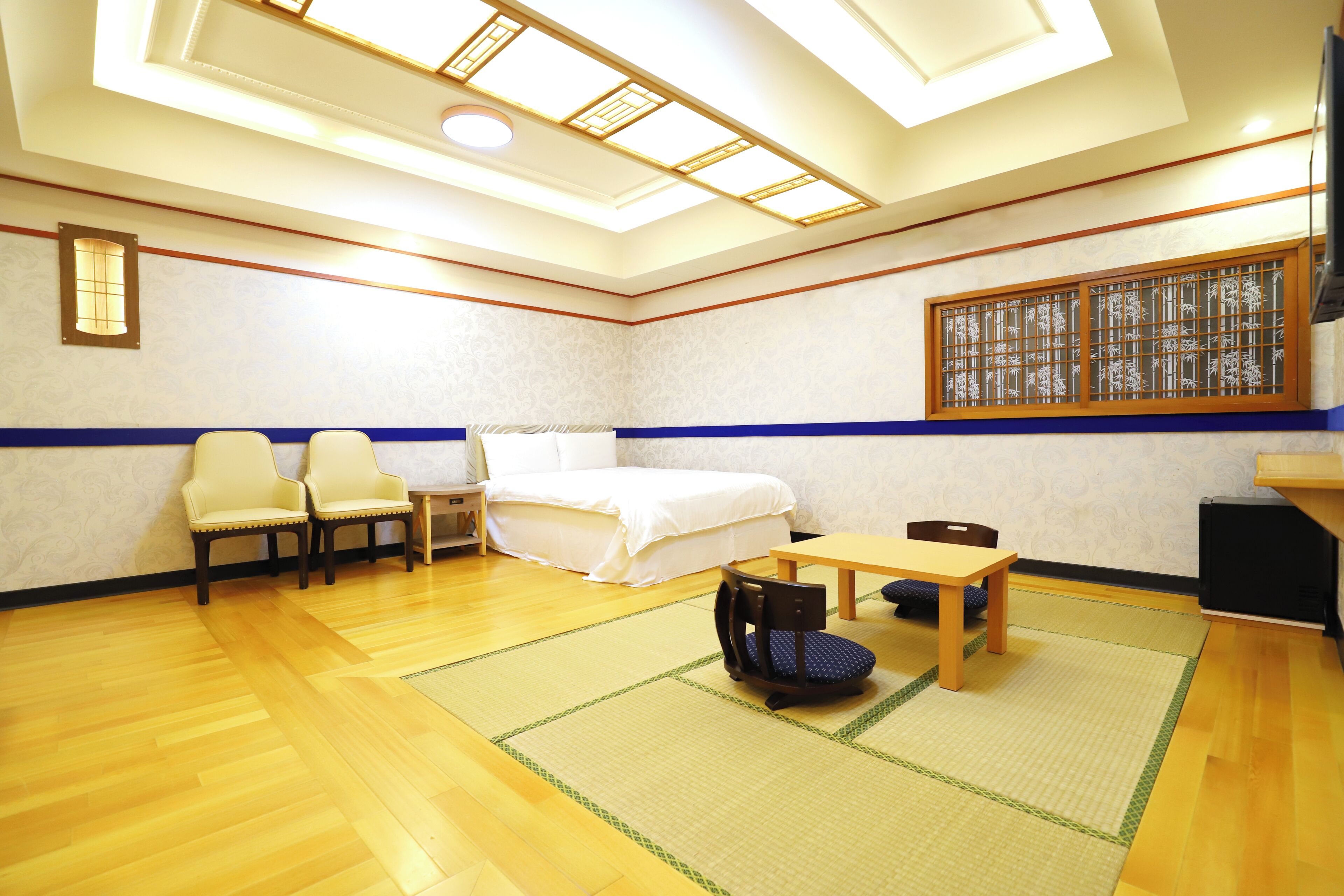 Habitació amb un llit, dues cadires, una taula petita, un matalàs de tatami, una televisió i una finestra amb un panell decoratiu.