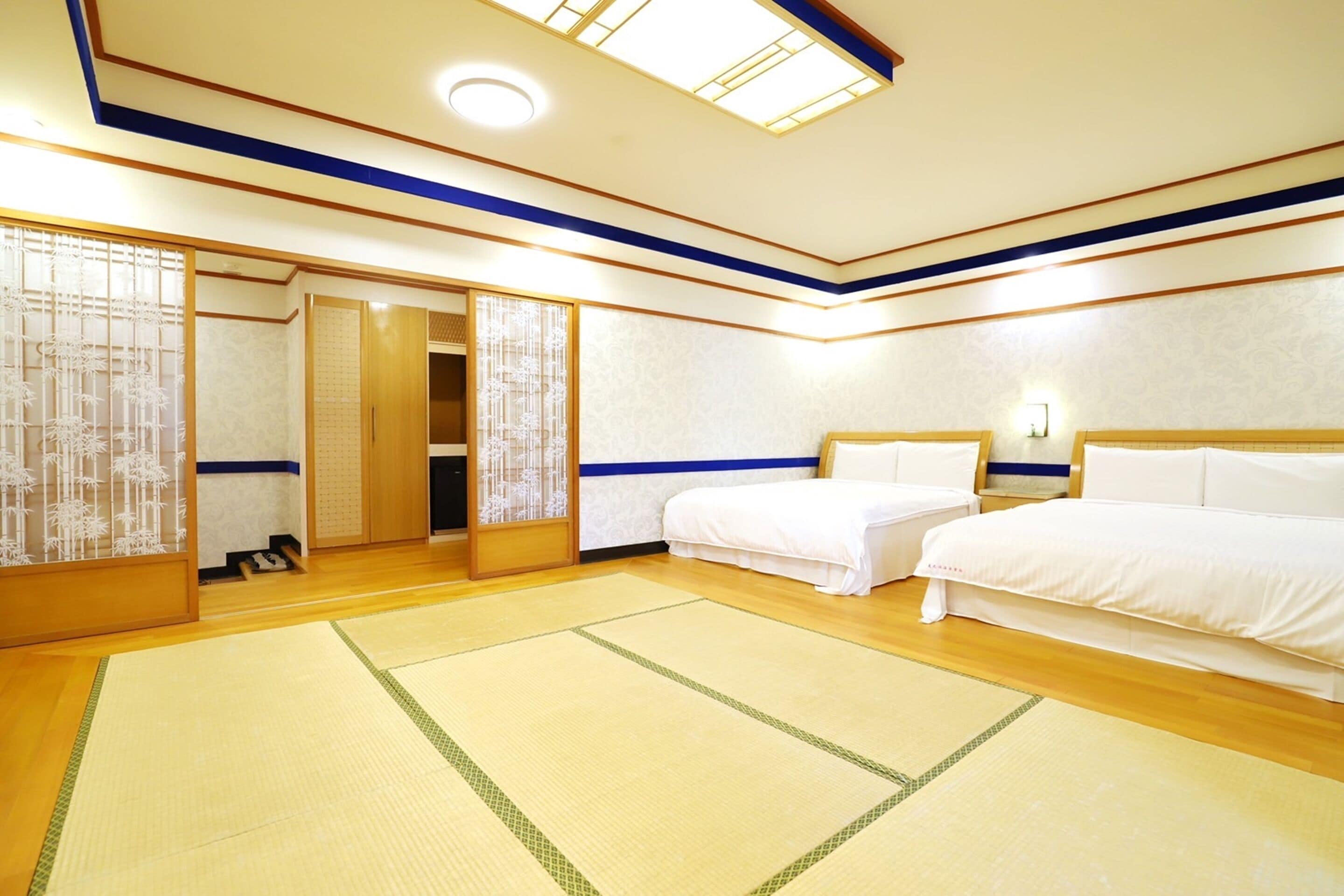 A hotel room with two beds, a wooden floor, tatami mats, a sliding door, and a ceiling light.