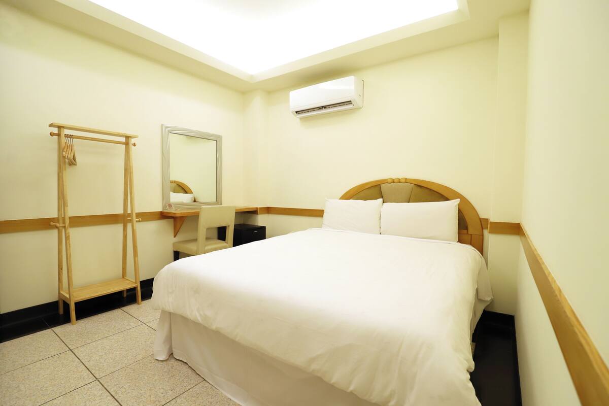A hotel room with a bed, a wooden ladder, a mirror, a desk, and an air conditioning unit.