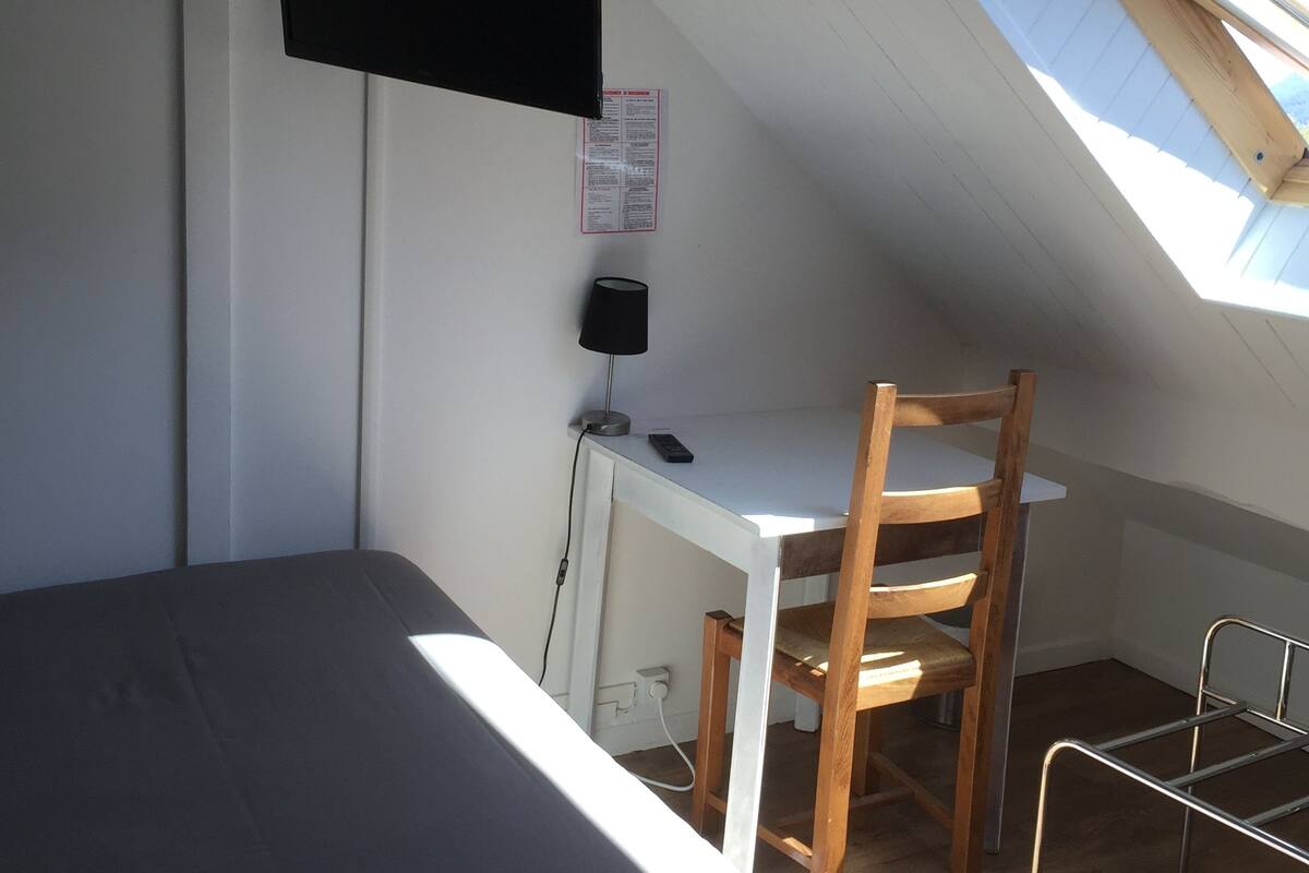 A small, slanted room with a bed, a desk, a chair, and a TV mounted on the wall.