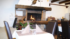 A dining area with a brick fireplace, wooden beams, and a table set for two with a white tablecloth. - Auberge de Violaine (Monetier-les-Bains)