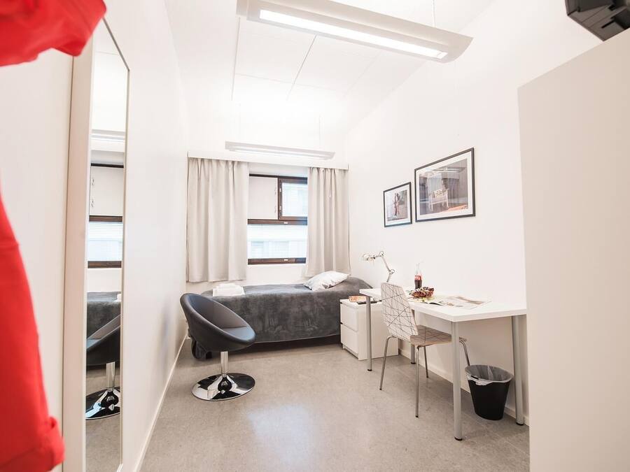 Habitación de hotel pequeña con cama, escritorio, silla, cesto de basura y ventana con cortinas.