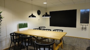 Una sala de conferencias con una larga mesa de madera, sillas negras, dos lámparas colgantes, un reloj, una pizarra y una pantalla para proyector.