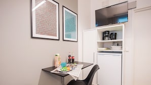 Una kitchenette compacta con refrigerador, microondas y cafetera. Hay una pequeña mesa de comedor con una silla, una cama y dos cuadros en la pared.