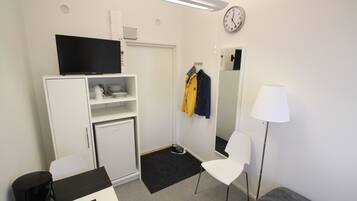 A compact room with a flat-screen TV on a cabinet, a small table with a coffee maker, a white chair, a sofa, a mirror, and a clock on the wall.