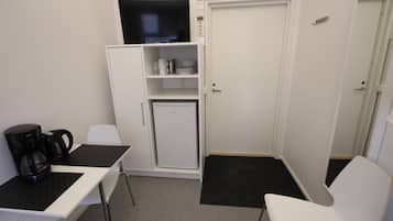 A compact room with a white desk, a coffee maker, a mini-fridge, and a television on a shelf.