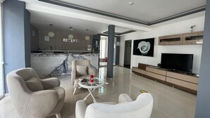 Un salon moderne avec un canapé, un fauteuil et une table basse. On aperçoit un téléviseur posé sur un meuble, un pare-feu en marbre et, au fond, un comptoir d’accueil.
