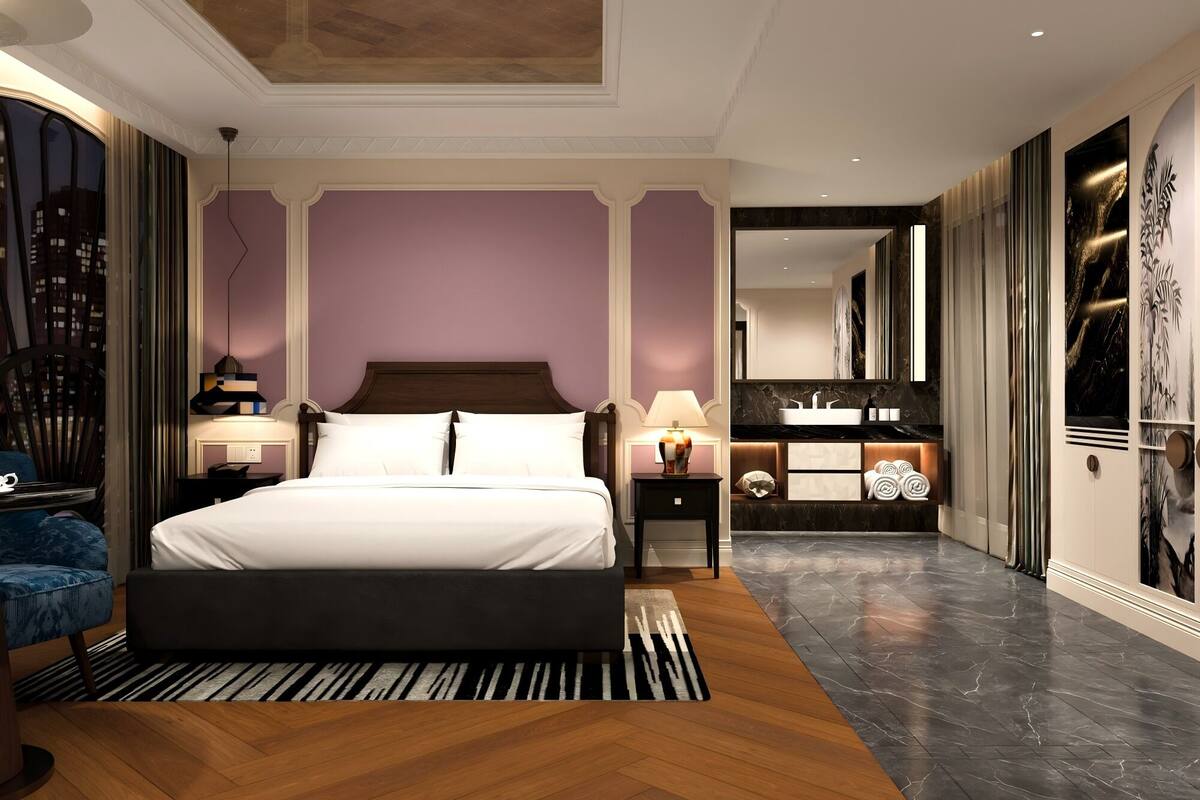A modern hotel room with a large bed, a sitting area with a chair, a vanity with a mirror, and a view of the city.