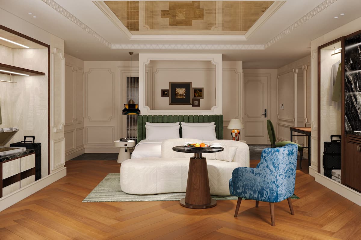 A spacious hotel room with a large bed, a sitting area with a chair, a small round table, and a desk with a lamp.