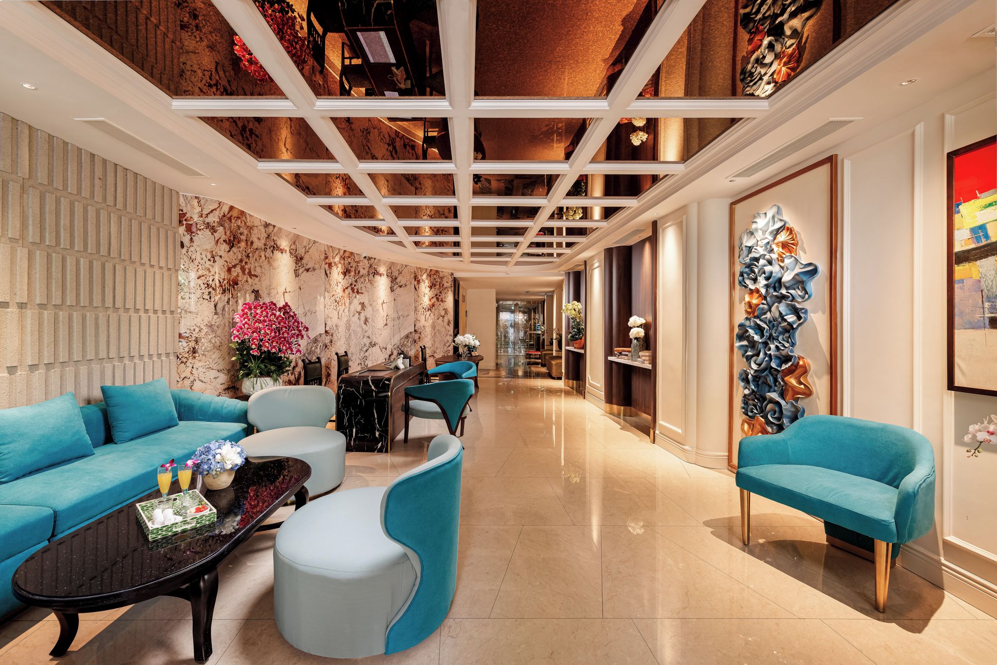 A modern hotel lobby with a marble wall, a seating area with blue sofas, a coffee table, and a decorative wall piece.
