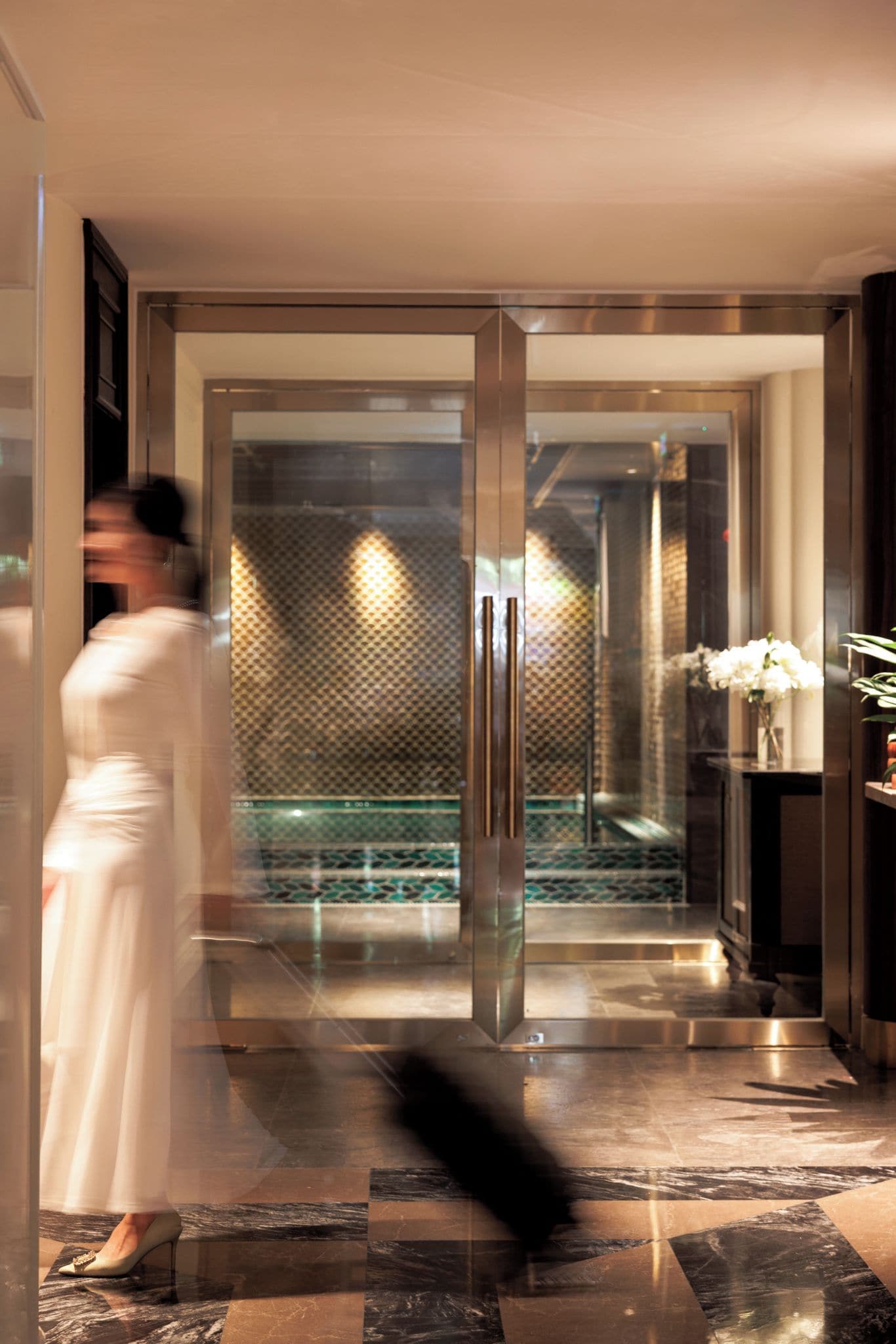 A hotel lobby with a glass door entrance, a marble floor, and a vase with white flowers on a table.