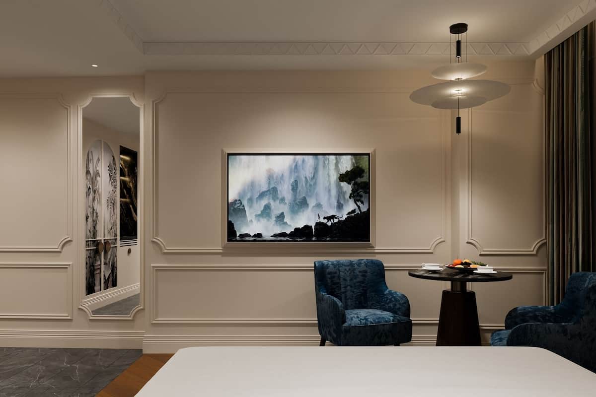 A hotel room with a large bed, two blue armchairs, a small table with a vase, and a large painting of a waterfall on the wall.