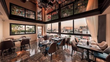 A dining area with multiple tables and chairs, large windows offering a city view, and a ceiling with hanging lights.