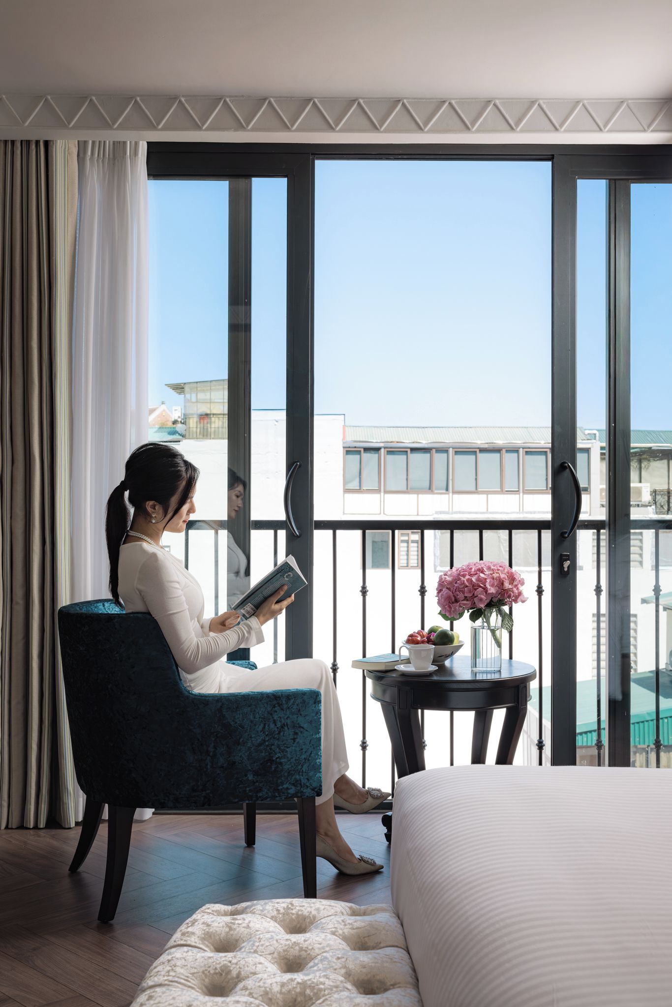 Une femme est assise dans une chambre d’hôtel, lisant près d’une grande fenêtre. On aperçoit une petite table sur laquelle est posé un vase de fleurs, ainsi qu’un lit recouvert d’une couette blanche.