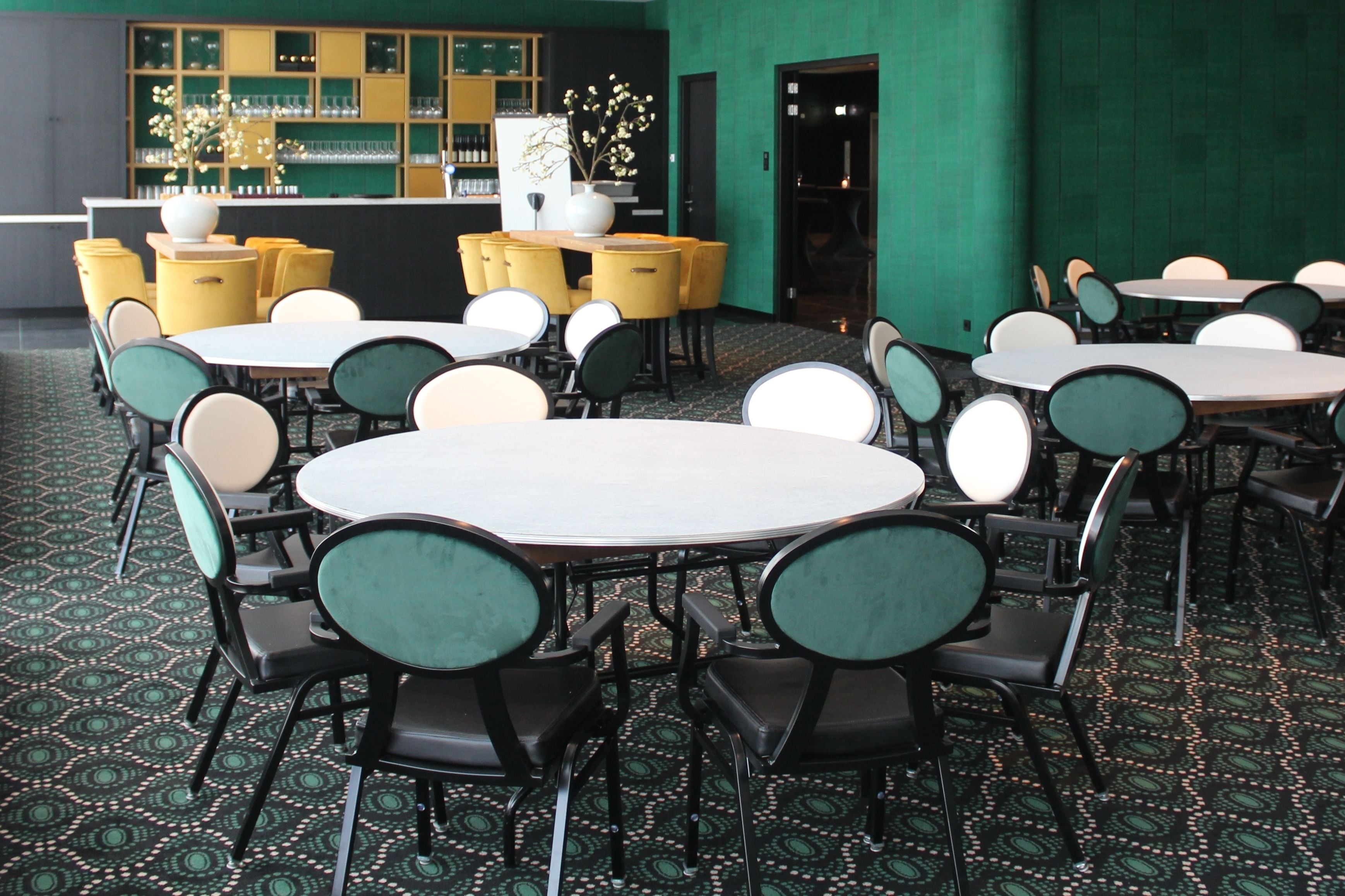 A restaurant with round tables and chairs, a bar area with shelves of glasses, and a patterned carpet.
