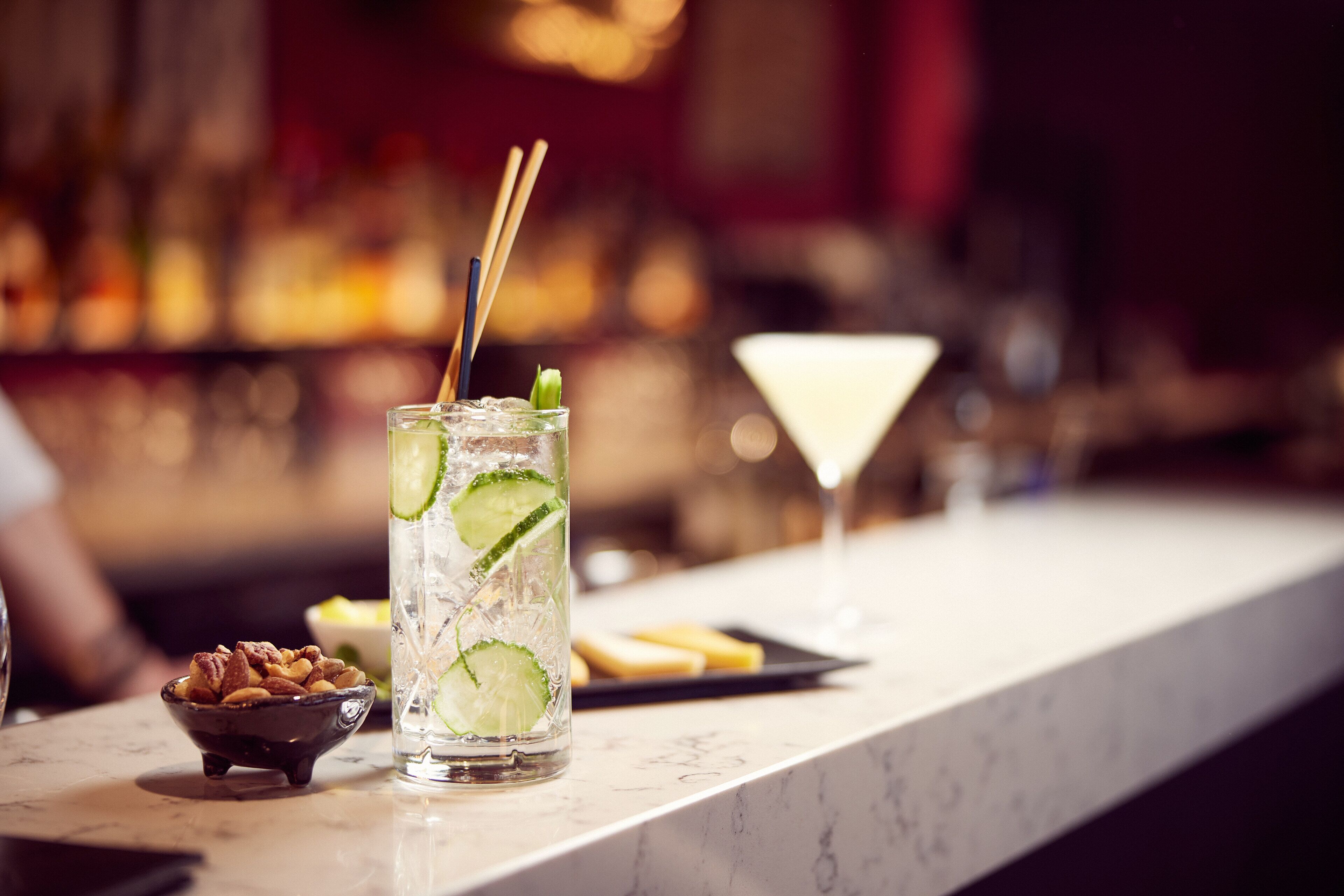 Un bar con un vaso de bebida a base de lima, una copa de martini y un cuenco pequeño de nueces sobre una encimera de mármol.
