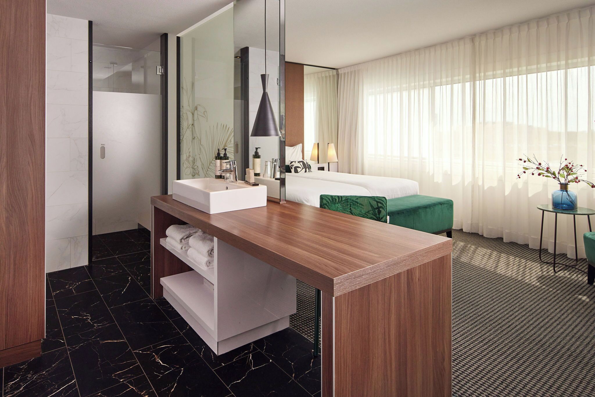 Habitación de hotel moderna con tocador de madera, un espejo grande, cama con ropa blanca, una butaca verde y vistas al exterior a través de cortinas traslúcidas.