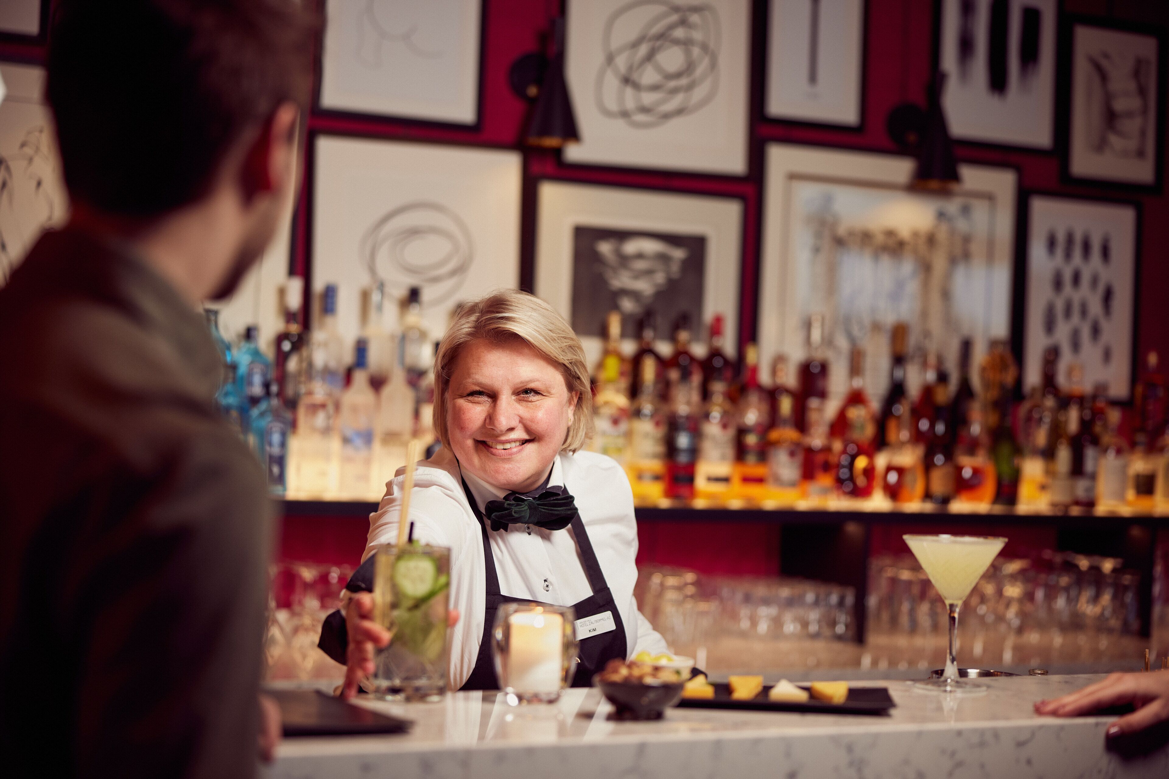 A bar with a bartender, a cocktail, and various bottles in the background.