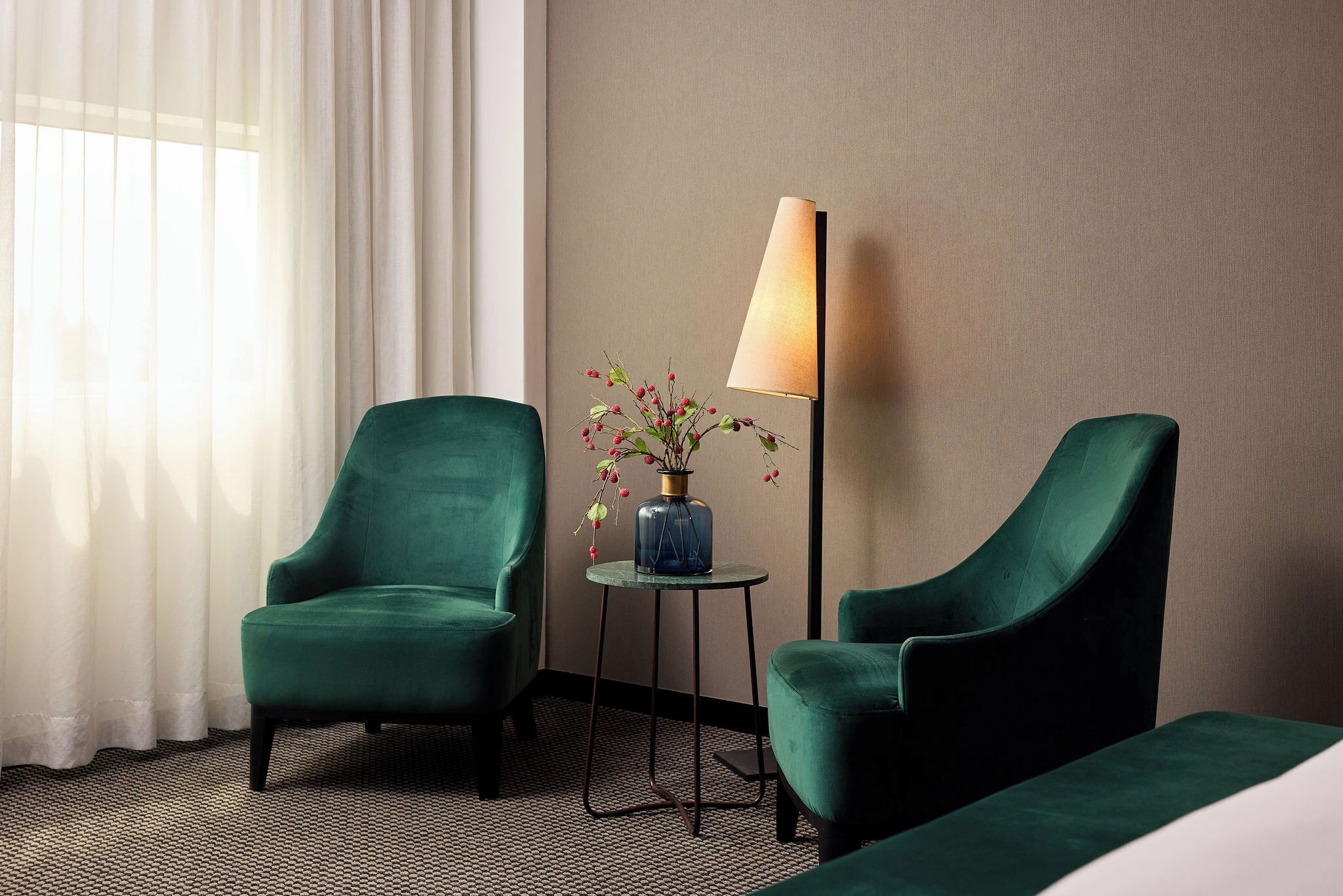 Een hotelkamer met twee groene fauteuils, een tafeltje met een bloevas en een lamp.