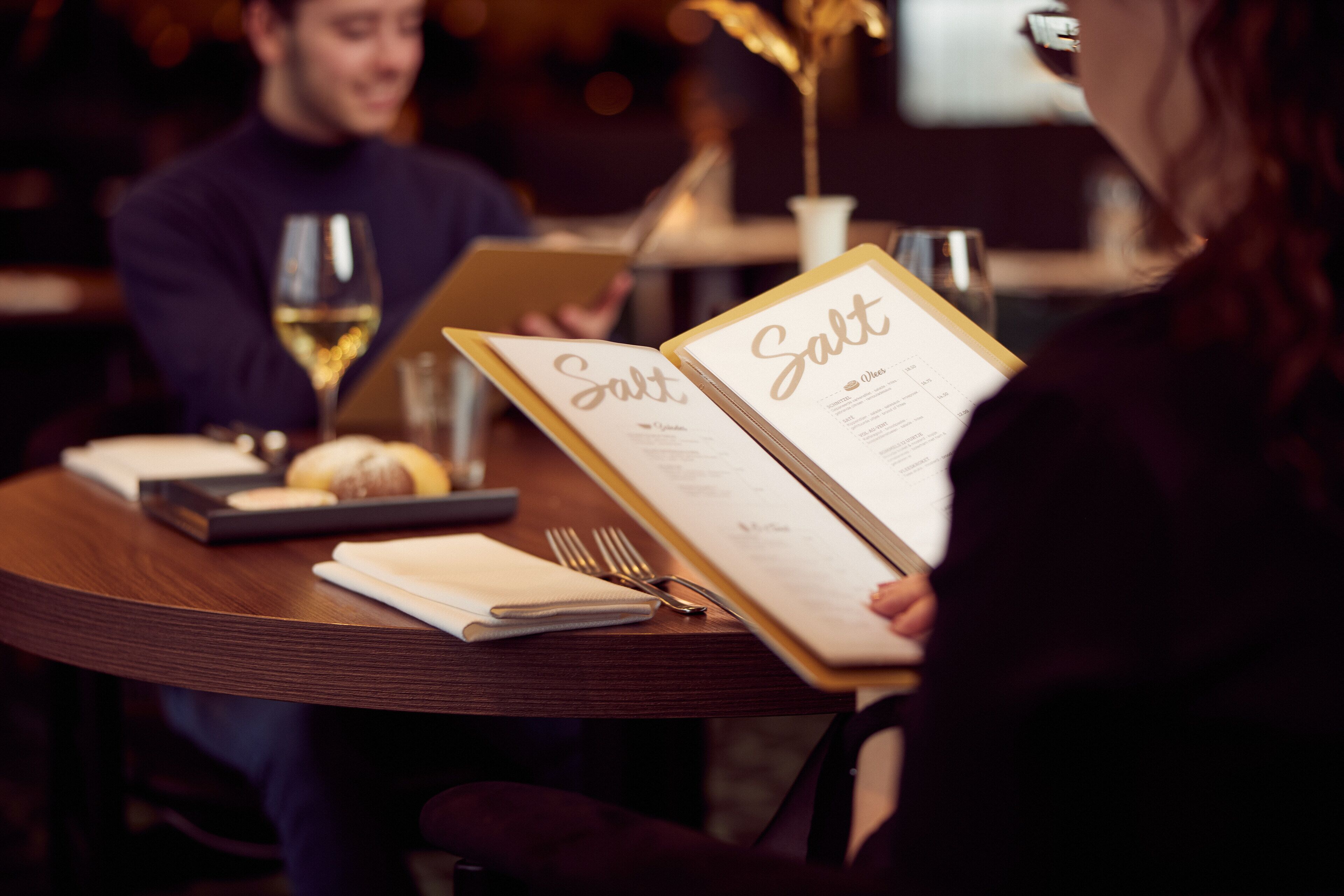 A person holding a menu with the word "Salt" on it, sitting at a wooden table with a laptop, wine glass, and bread.