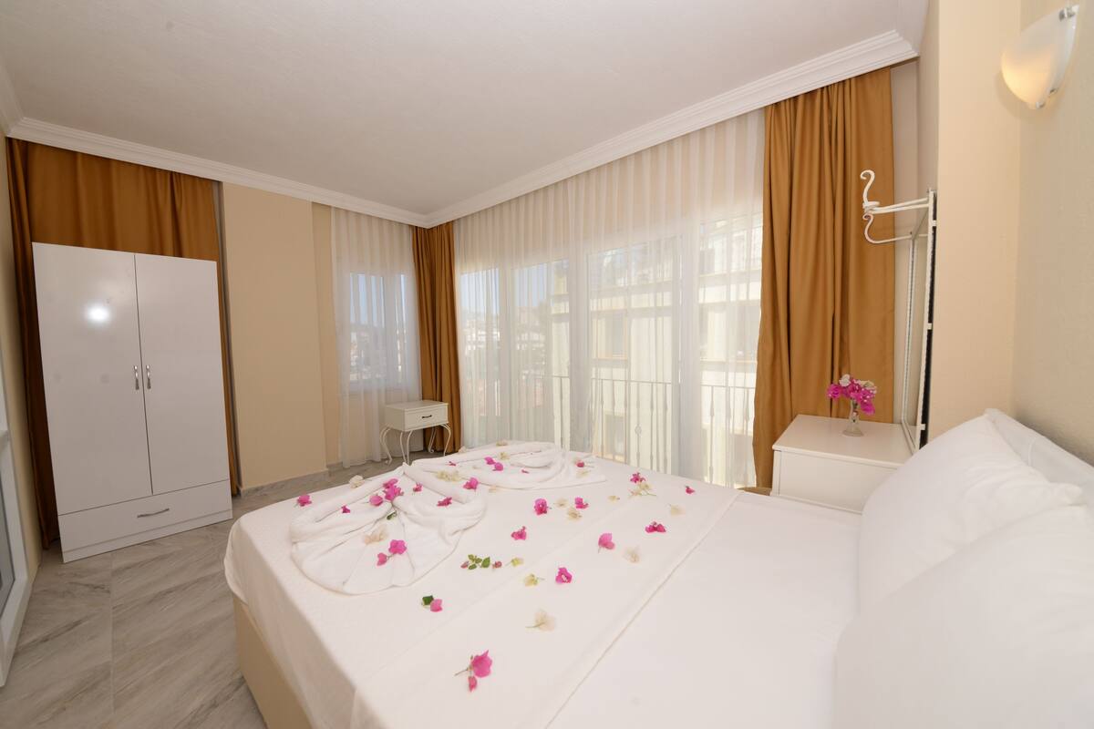 A hotel room with a large bed, a wardrobe, a bedside table with a vase of flowers, and a window with curtains.