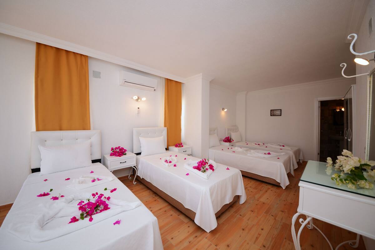 A hotel room with three single beds, each with a white bedspread and pink flowers. There are white nightstands with lamps, a wooden floor, and a glass table with a vase of flowers.