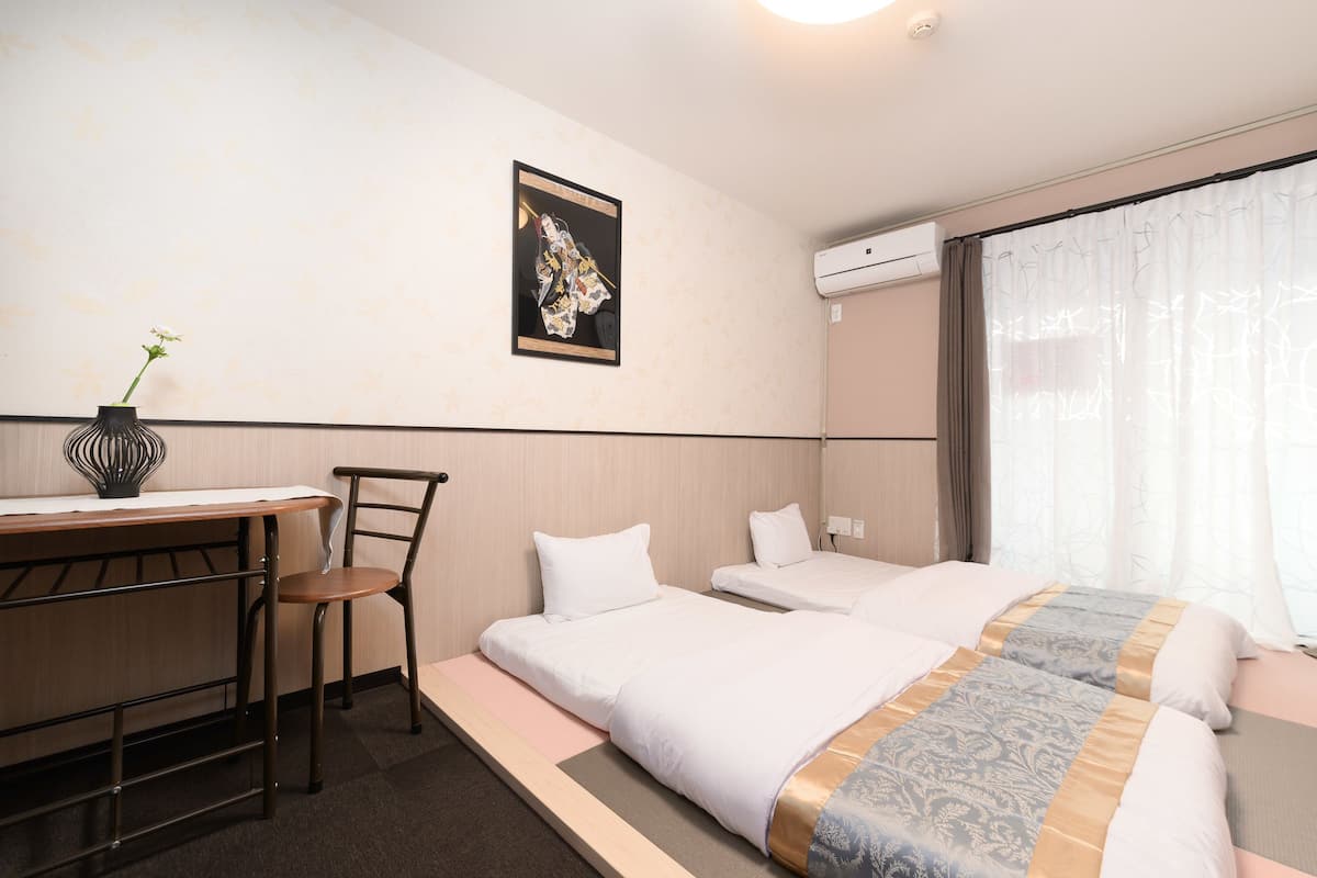 Quarto de hotel com escrivaninha, duas camas de solteiro, uma cadeira, ar condicionado fixado na parede e uma janela com cortinas.