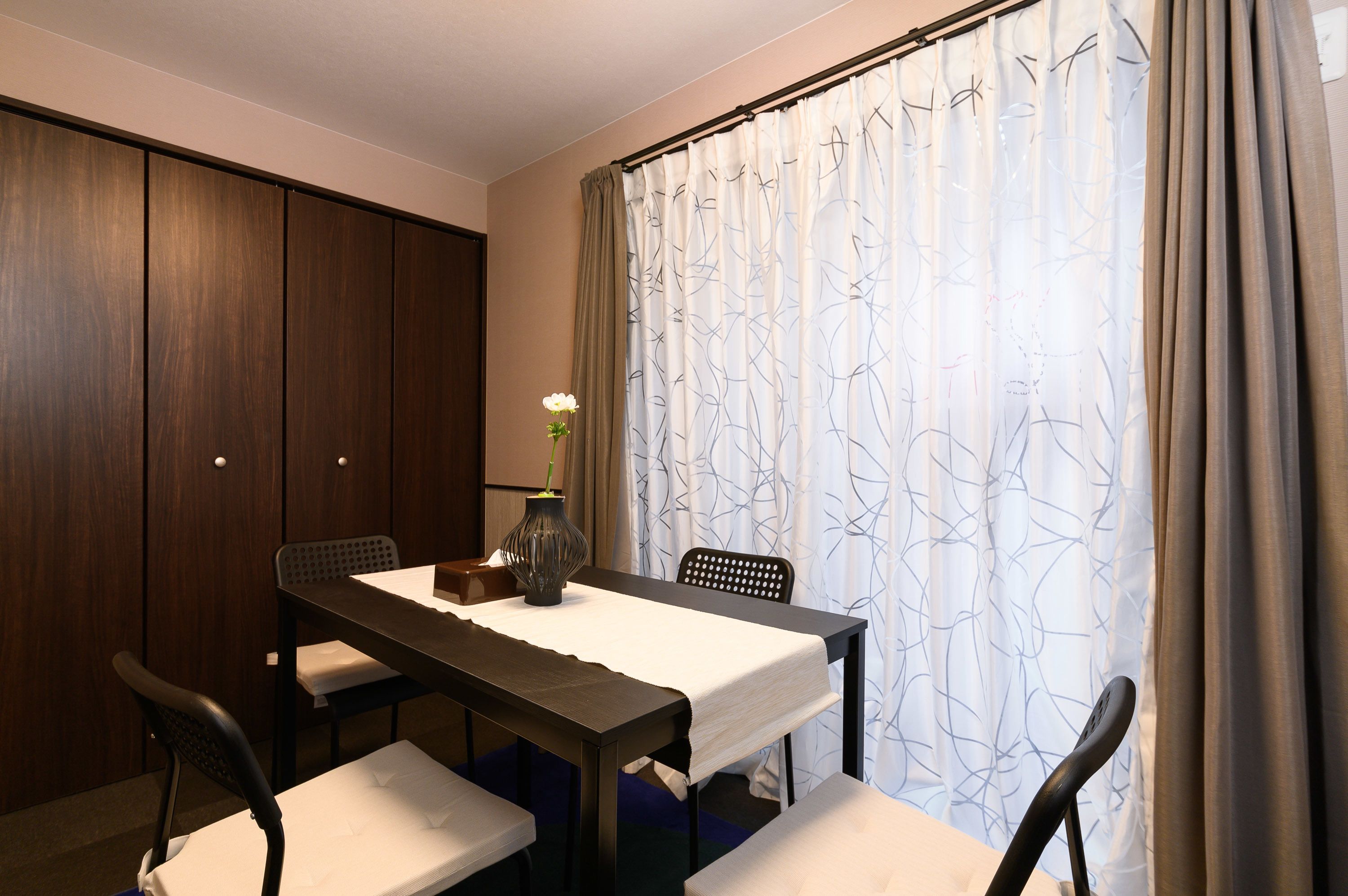 A dining area with a table set for four, a vase with flowers, and a view through patterned curtains.