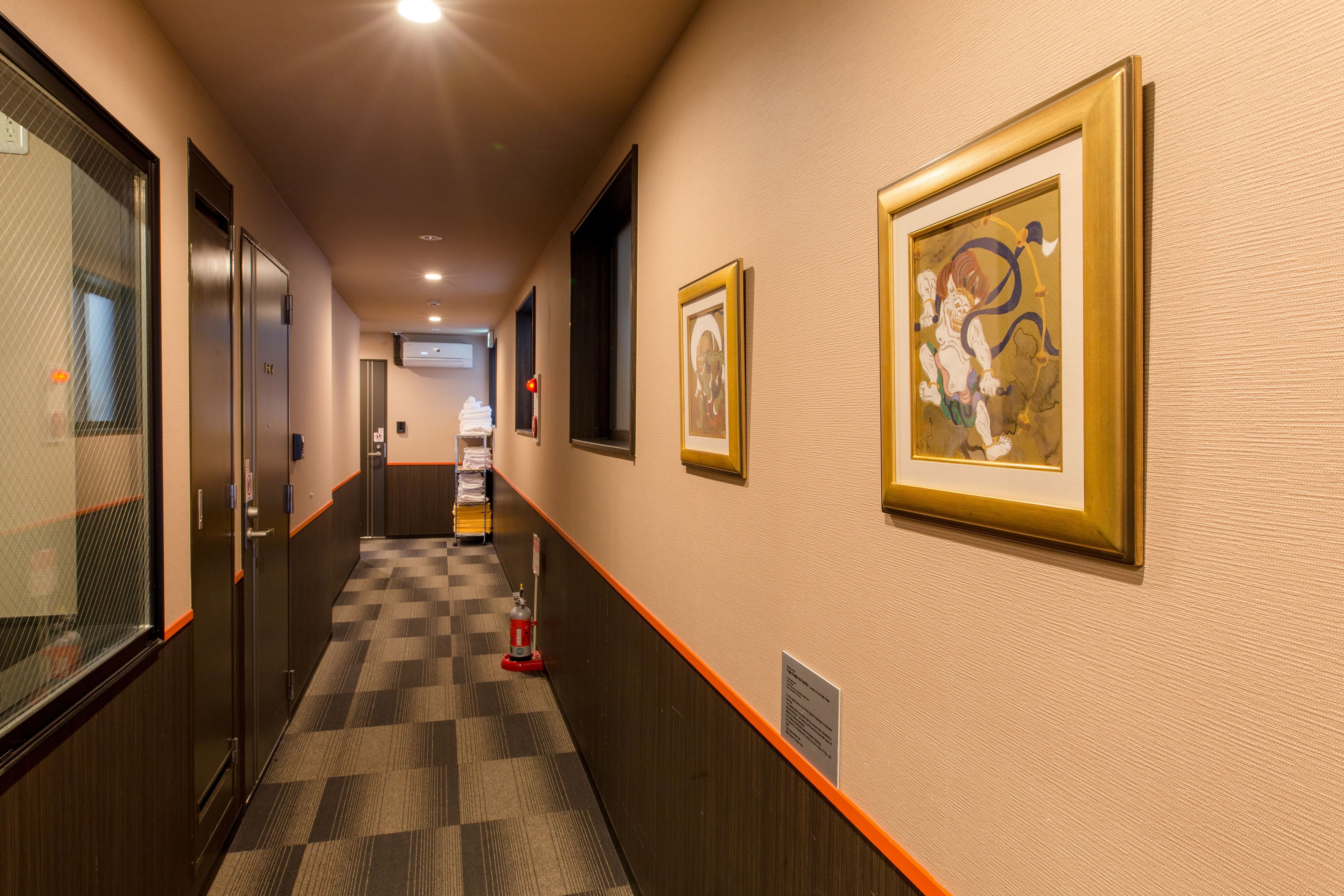 Een hotelgang met een geruit tapijt, kunstwerken in lijsten aan de muren en een brandblusser op de vloer.