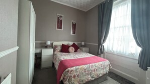 Quarto com cama, mesas de cabeceira, cômoda, janela com cortinas e duas imagens penduradas na parede.