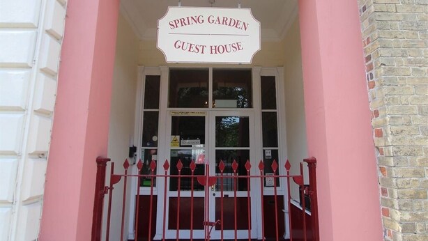 Entrada da Guest House Spring Garden, com uma placa branca sobre a porta e um portão vermelho.