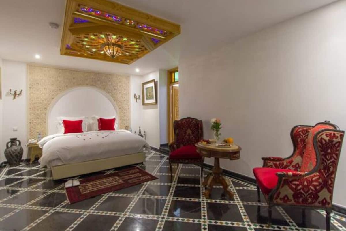 A hotel room with a bed, a red chair, a small table with a vase, and a decorative ceiling light.