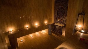 Zona de spa tenuemente iluminada con un banco de madera, un lavamanos pequeño y una pared decorativa con diseños intrincados.