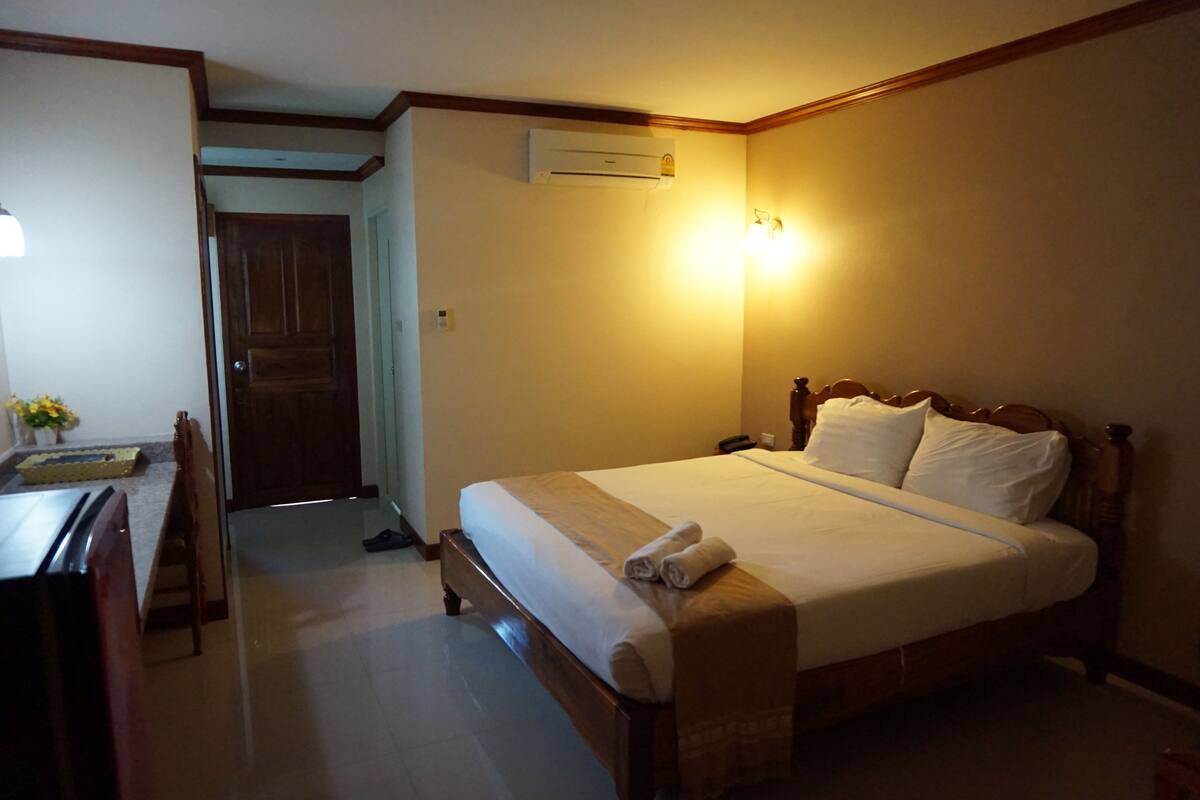Ein Hotelzimmer mit einem hölzernen Bett, einem Schreibtisch mit Stuhl, einer Klimaanlage und einer Tür zu einem anderen Raum.