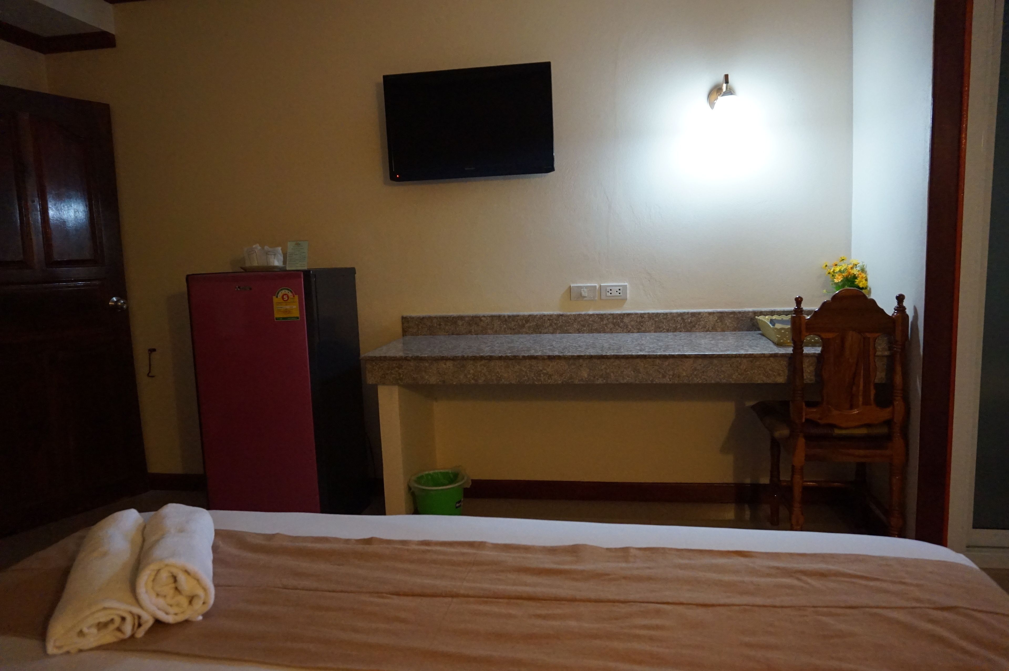 Una habitación de hotel con cama, un escritorio con silla, televisor montado en la pared y un refrigerador pequeño.