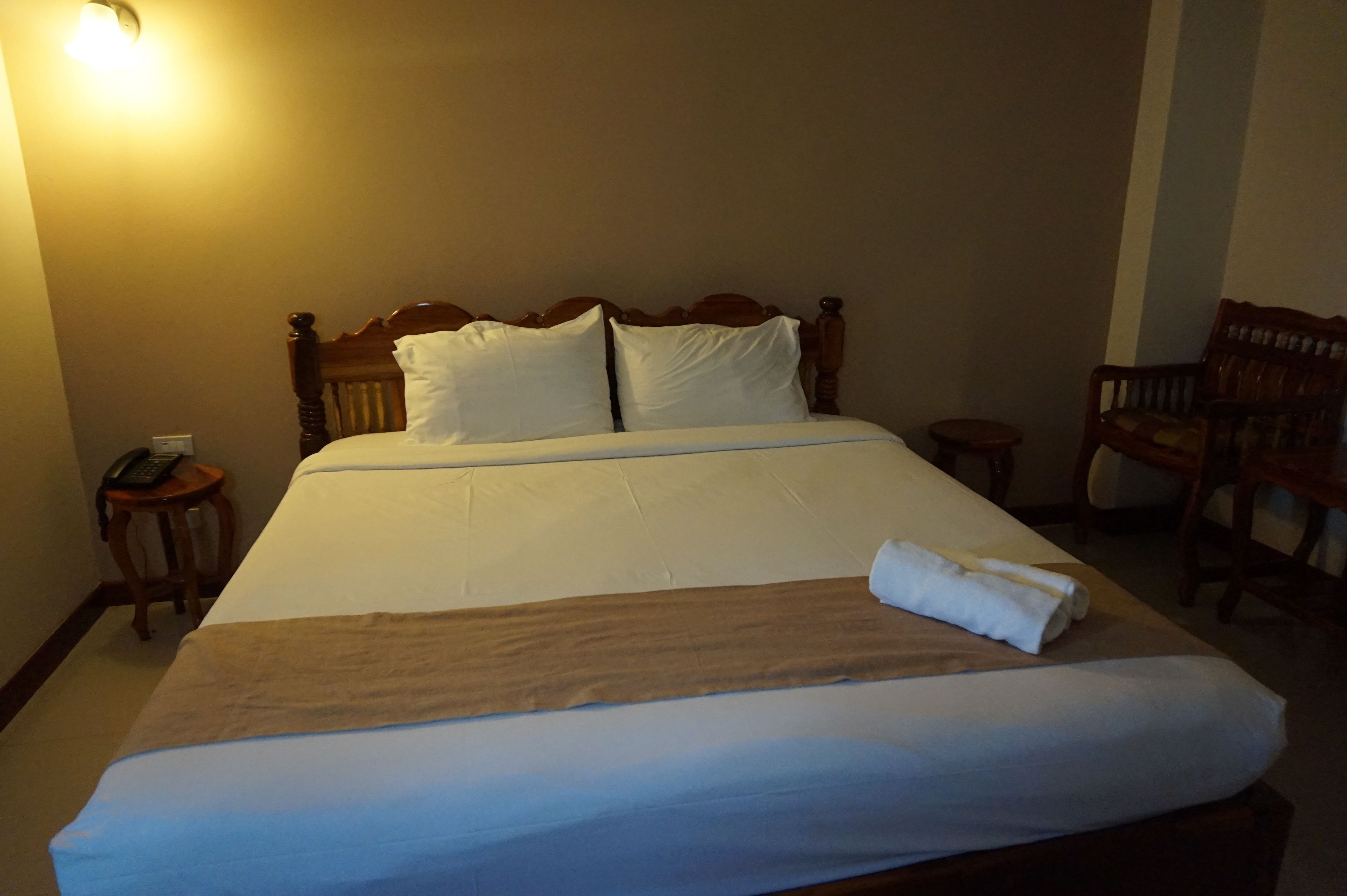 Una cama bien hecha con cabecera de madera, una mesita de noche con un teléfono y una silla a la derecha.