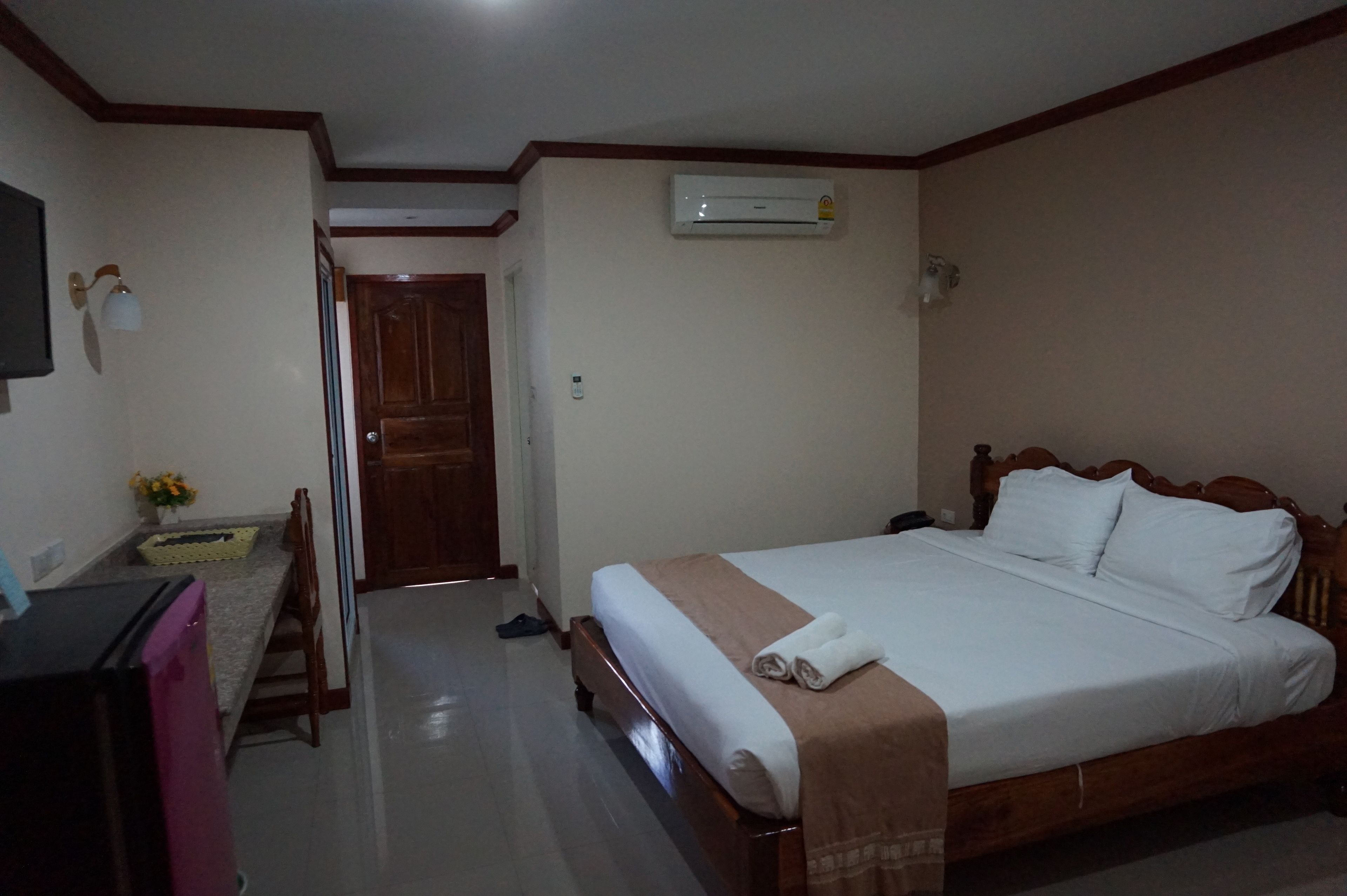 A hotel room with a bed, a desk, a chair, a small table with flowers, an air conditioning unit, and a door.
