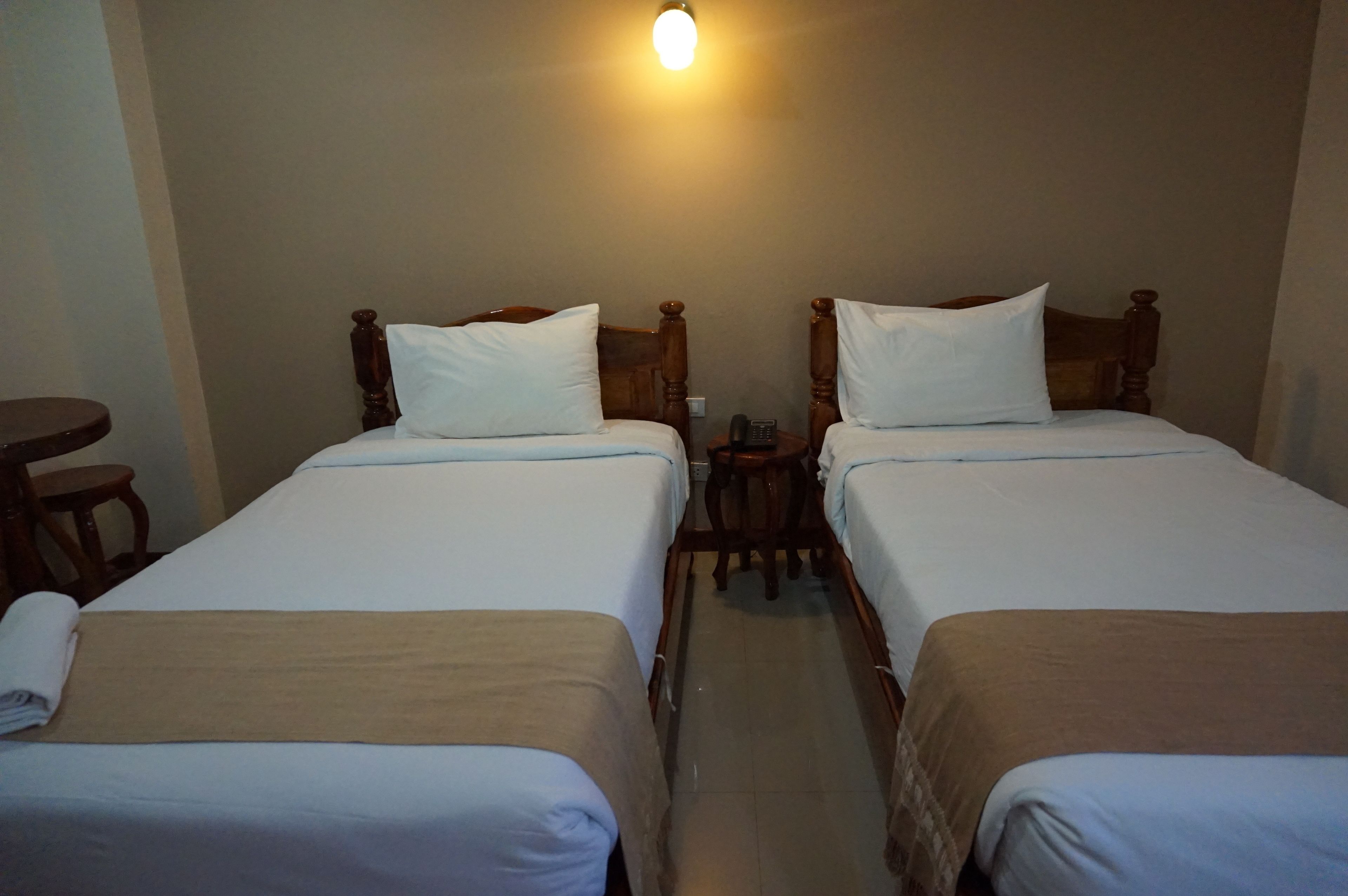 A room with two single beds, each with a wooden headboard, a small bedside table, and a wall-mounted light fixture.