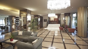 A spacious lobby with a grand chandelier, a staircase leading to a dining area, and a bar with a wine collection. - Hotel Calissano (Alba)