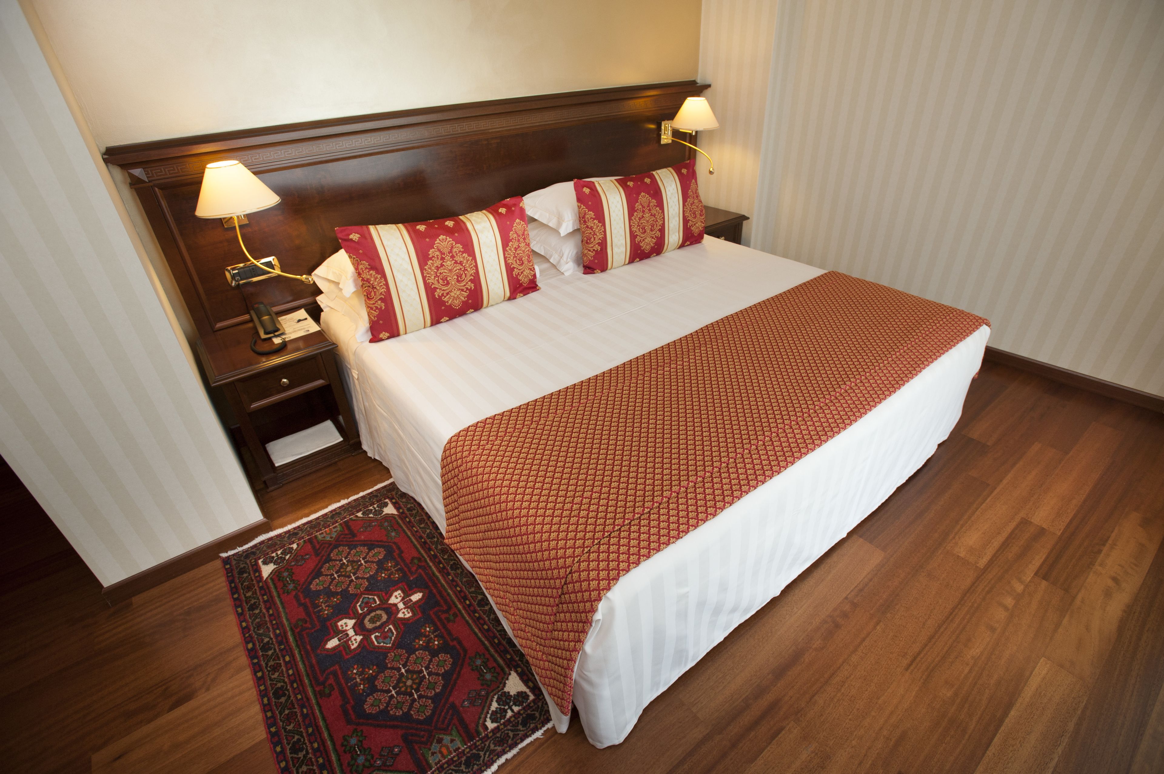 Una cama bien hecha con una colcha roja estampada, dos almohadas con diseños rojo y blanco, un cabecero de madera, una mesita de noche con una lámpara y una alfombra roja y negra con estampado.