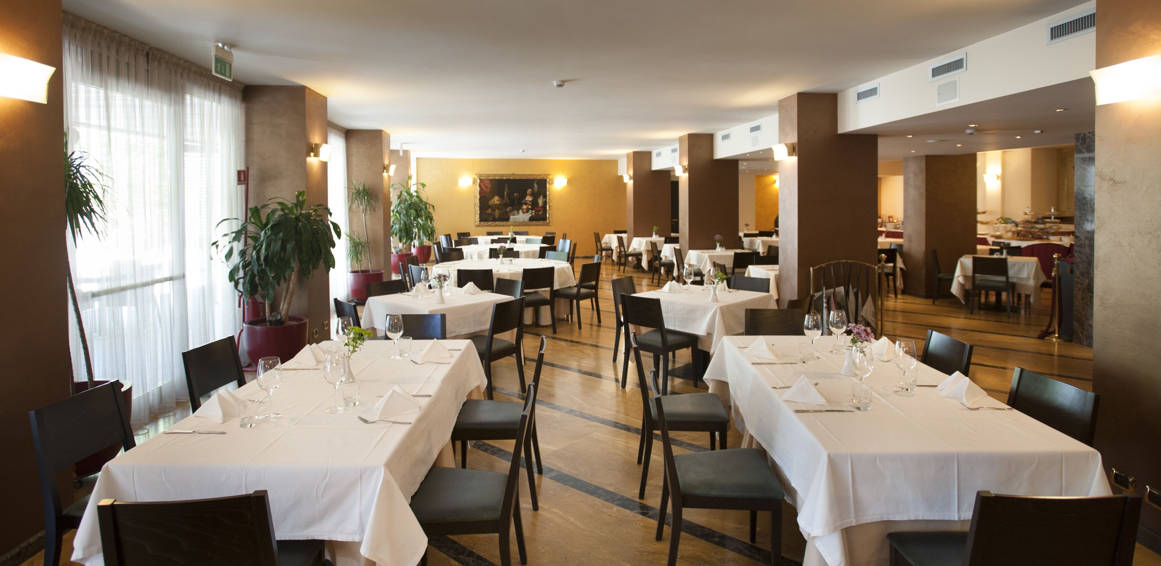 A hotel dining area with multiple tables set for dining, chairs, and potted plants.