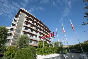 A multi-story hotel with a prominent sign, surrounded by greenery and flags. - Hotel Calissano (Alba)