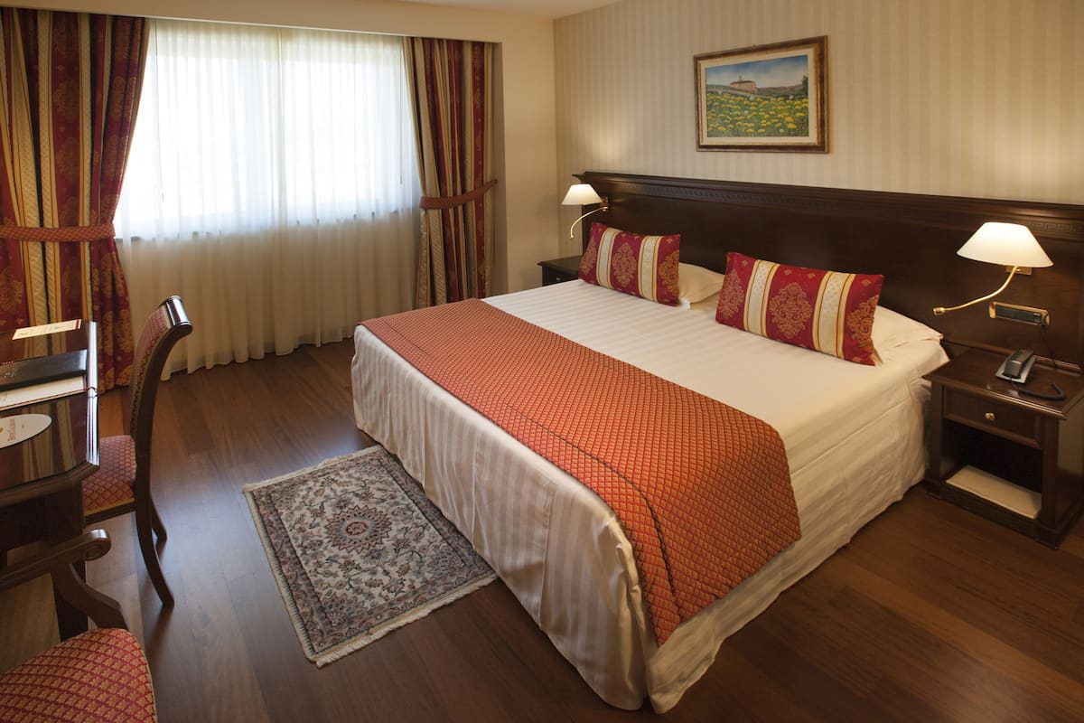Habitación de hotel con una cama grande, dos mesitas de noche con lámparas, una silla, un escritorio y un ventanal con cortinas.