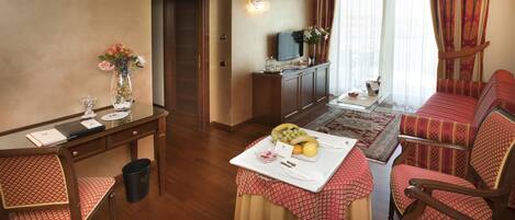A hotel room with a wooden desk, a TV, a dining table with fruit, and a sofa.