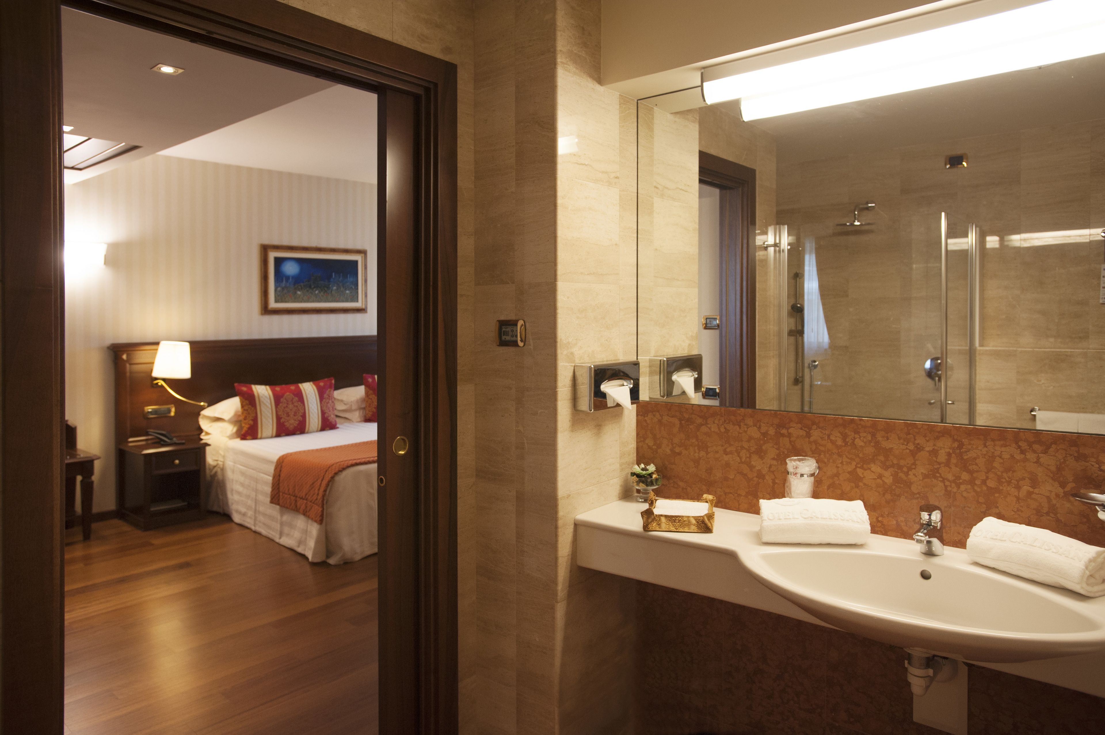A hotel room with a bed, bedside table, lamp, and a bathroom with a sink, mirror, and shower.