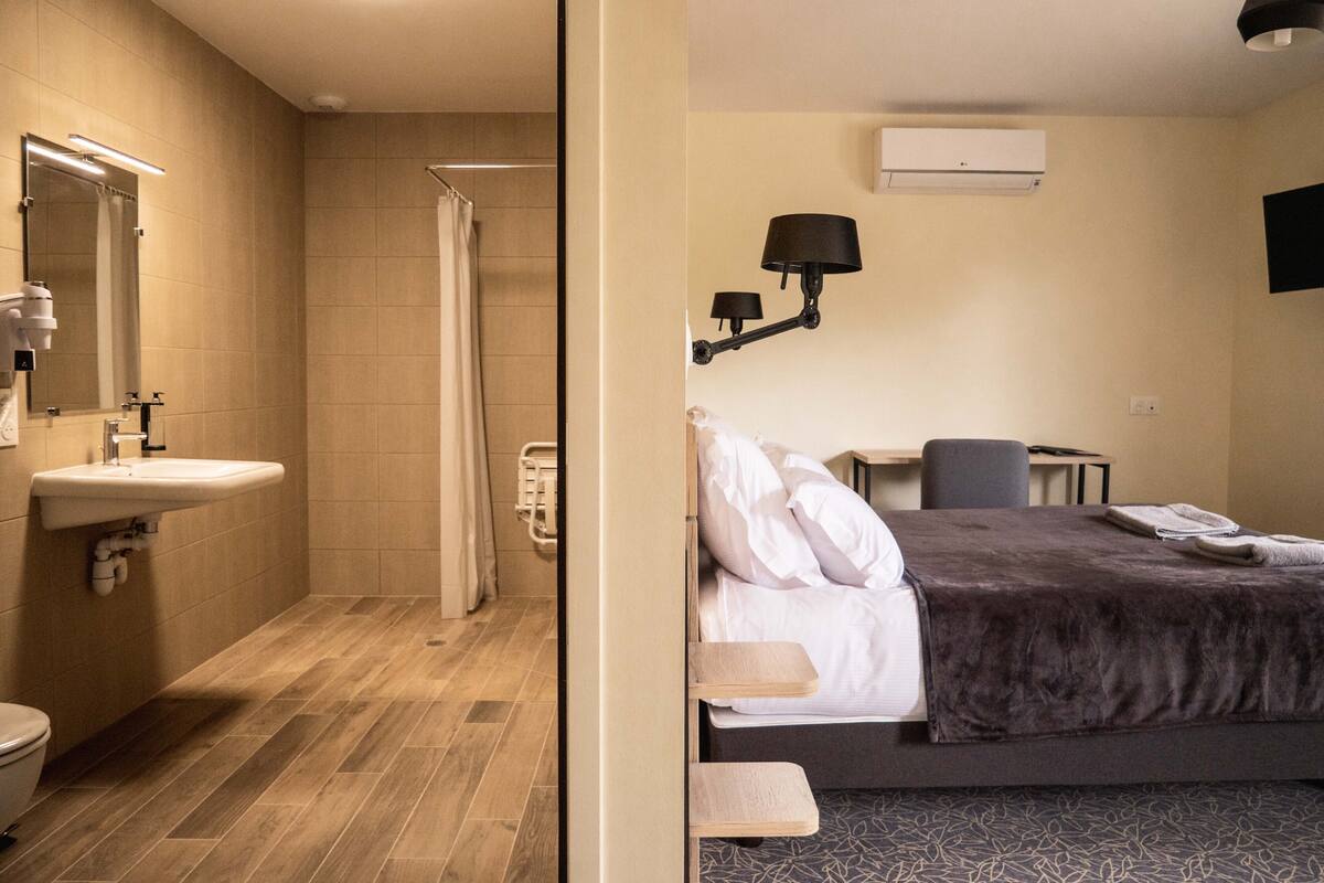 A hotel room with a shower, sink, bed, bedside table, and a TV mounted on the wall.