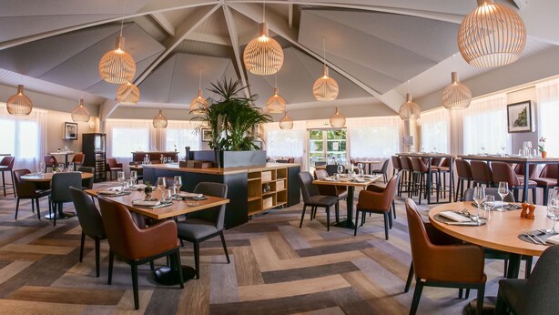 Un restaurante moderno con techo alto y diseño geométrico, que cuenta con una isla central y varias zonas de comedor con mesas y sillas redondas.