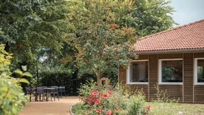 Una cabaña de madera con techo de tejas, rodeada por un jardín con flores rojas y un camino empedrado que lleva a una mesa y sillas.
