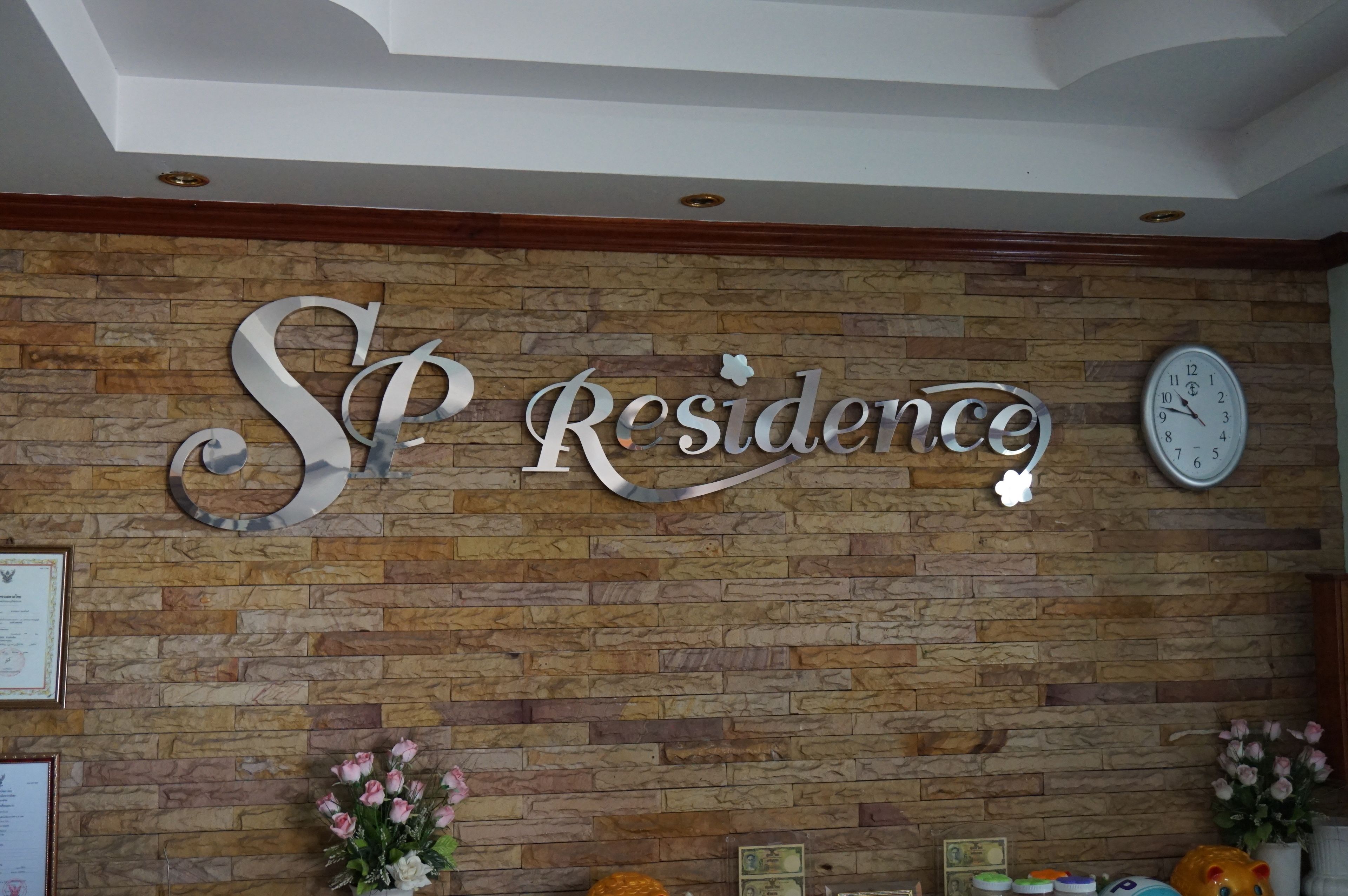 Un espace d’accueil avec un mur en pierre, une grande enseigne indiquant « SP Residence », une horloge et des décorations florales.